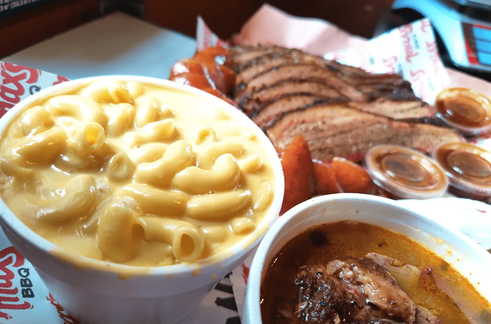 Vaso de mac n' cheese en primer plano con brisket rebanado y frijoles al fondo