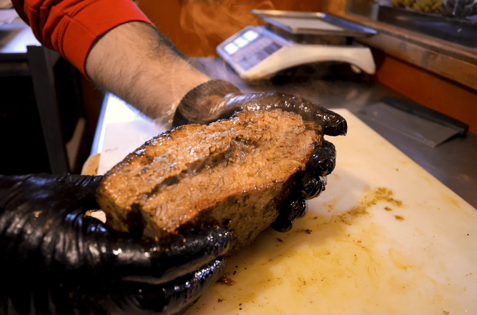Trozo de brisket humeante sostenido con guantes junto a la báscula del mostrador
